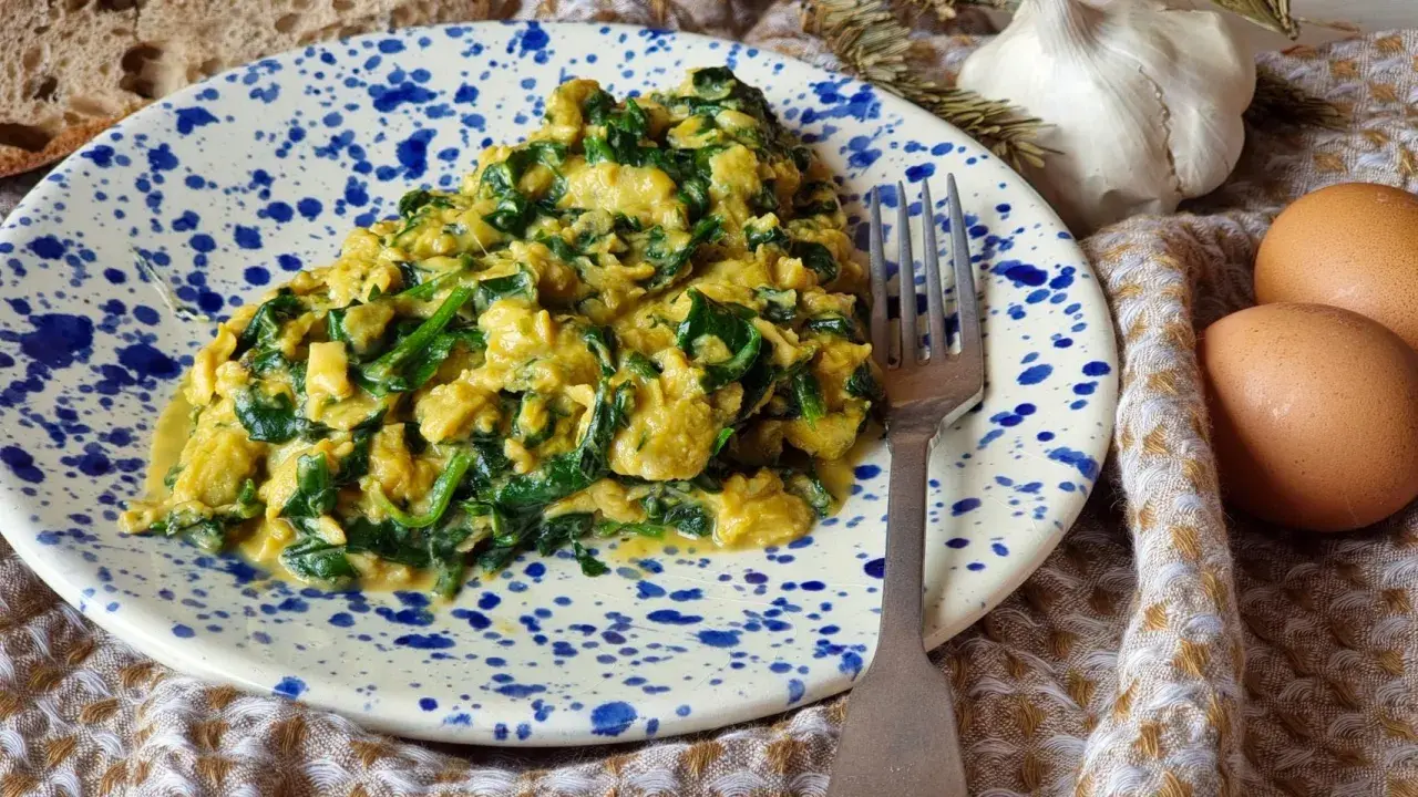 Un delicioso revuelto de espinacas cremoso, servido en un plato con salpicaduras azules. Acompañado de pan, ajo y huevos frescos.