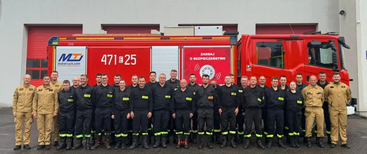 Grupa strażaków w mundurach, obok czerwonego wozu strażackiego. Dowódca sekcji OSP stoi w pierwszym rzędzie, w jasnym mundurze.