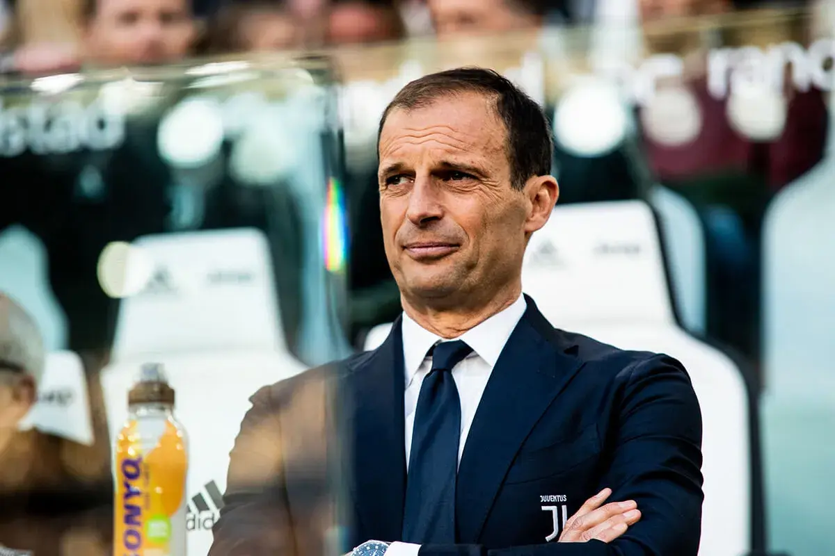 Massimiliano Allegri, w eleganckim garniturze, z rękami skrzyżowanymi na piersi, obserwuje grę.