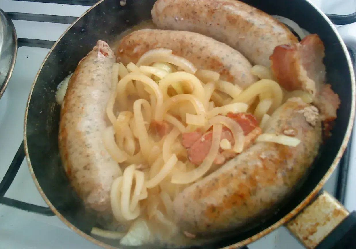 Biała kiełbasa w piwie dusi się z cebulą i boczkiem na patelni.