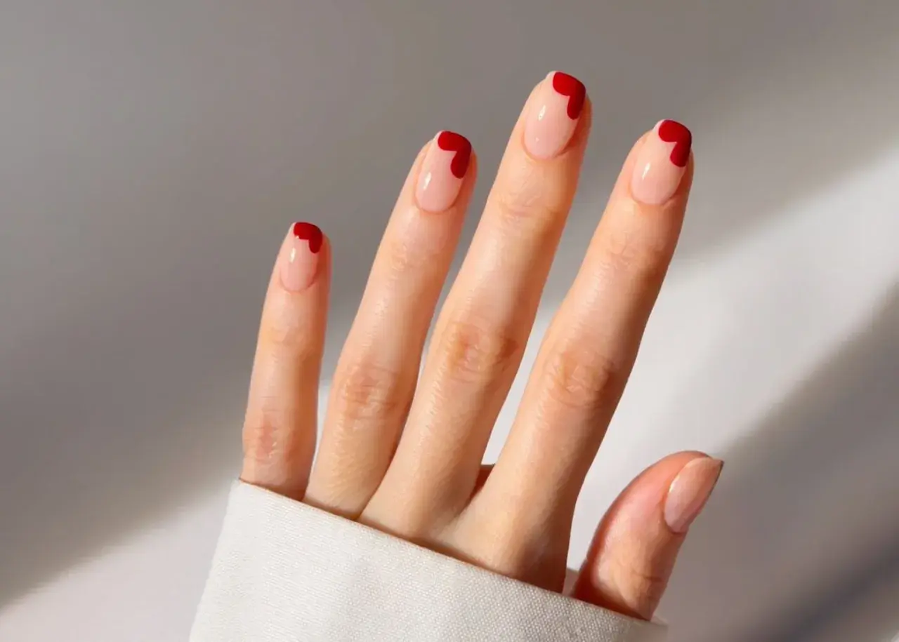 Une main aux ongles nude ornés de petits cœurs rouges. Une idée ongle coeur parfaite pour la Saint-Valentin ou pour ajouter une touche d'amour à votre quotidien.