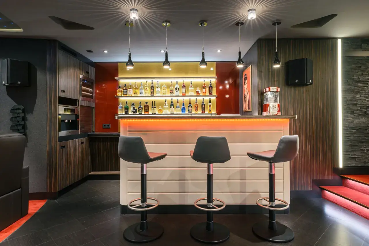 Stilvolle Bar mit beleuchteten Regalen voller Flaschen, drei Barhockern und einer Popcornmaschine. Tolle partyraum bar ideen für Ihr Zuhause.
