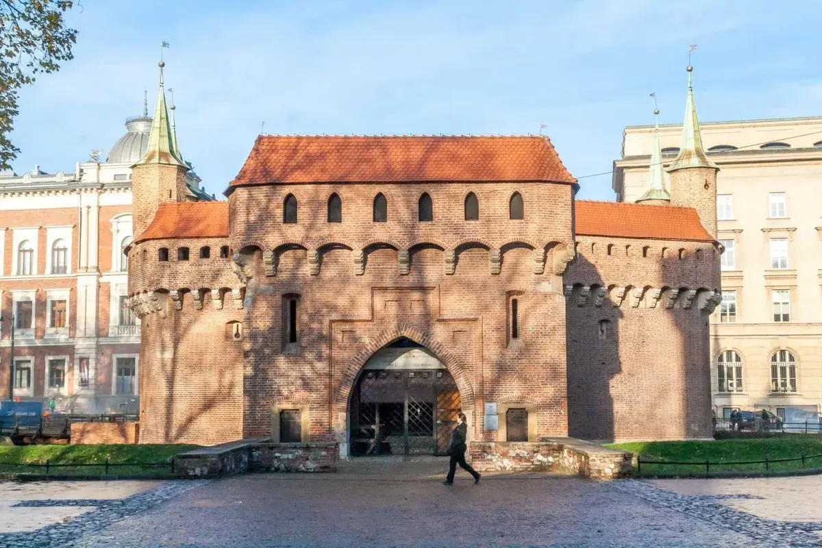 Krakowski barbakan z cegły, z charakterystycznymi wieżyczkami i łukowym przejściem, na tle błękitnego nieba.