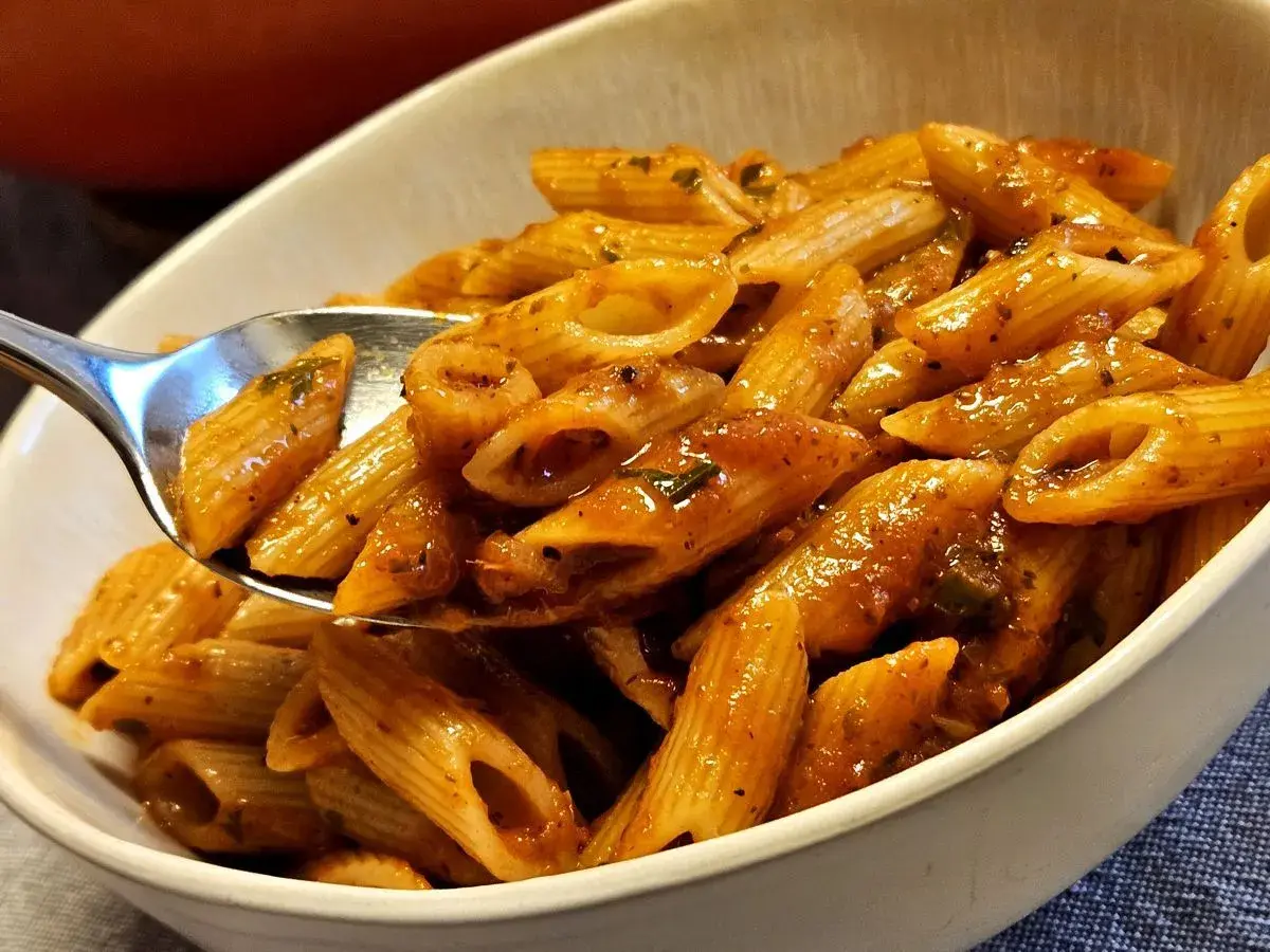 Eine Löffelportion Penne all'Arrabiata, mit einer würzigen Tomatensauce und Kräutern, wird aus einer Schüssel gehoben.