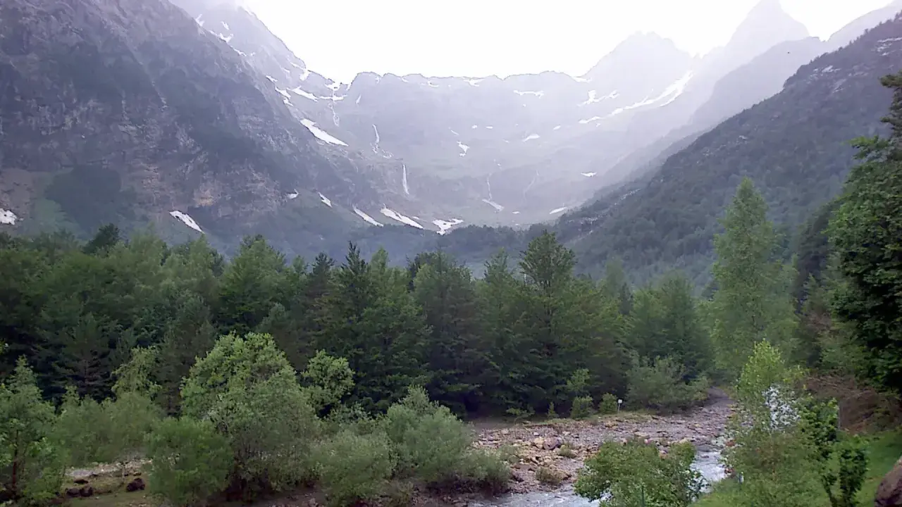 Majestuoso valle Pineta con cascadas cayendo por montañas cubiertas de nieve y un río serpenteando entre frondosos bosques verdes.