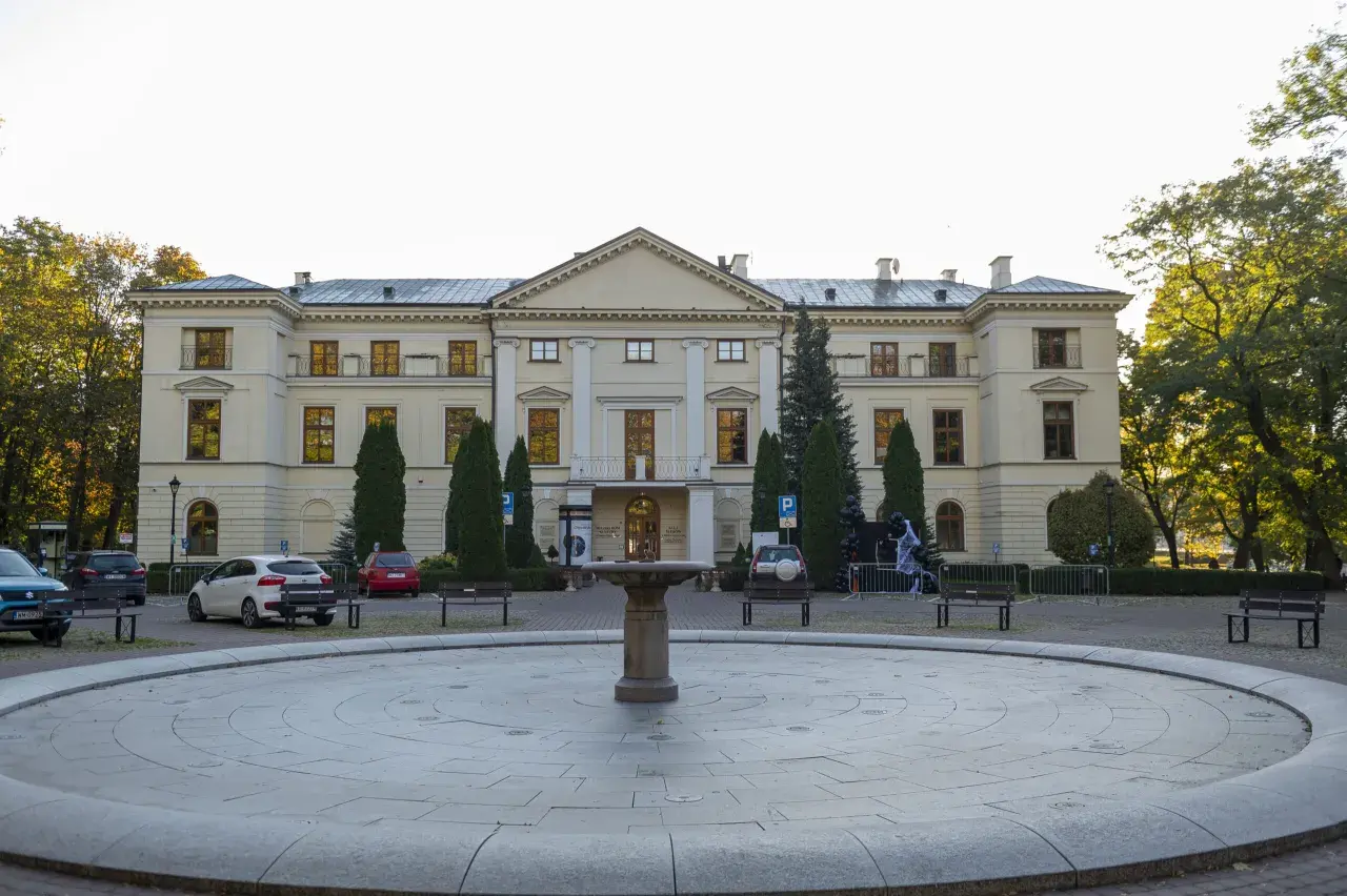 Pałac Dernalowiczów z fontanną i ławkami przed fasadą. W tle drzewa i zaparkowane samochody.