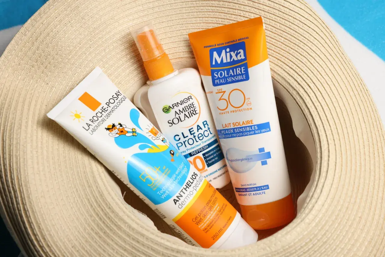 Trois tubes de crème solaire, dont une crème solaire périmée, sont posés sur un chapeau de paille.