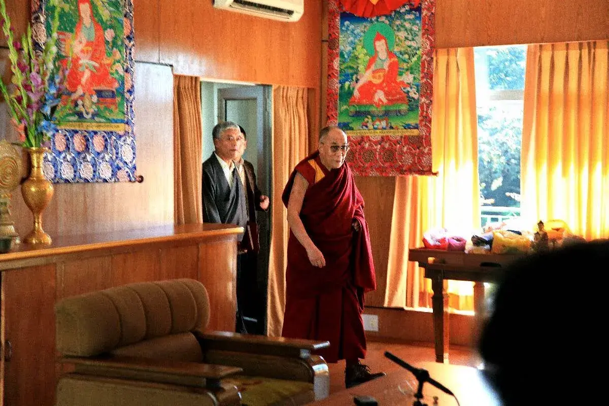 Dalai Lama i sin klädsel, med en man i kostym bakom. En inblick i var bor Dalai Lama, med buddhistisk konst på väggarna.