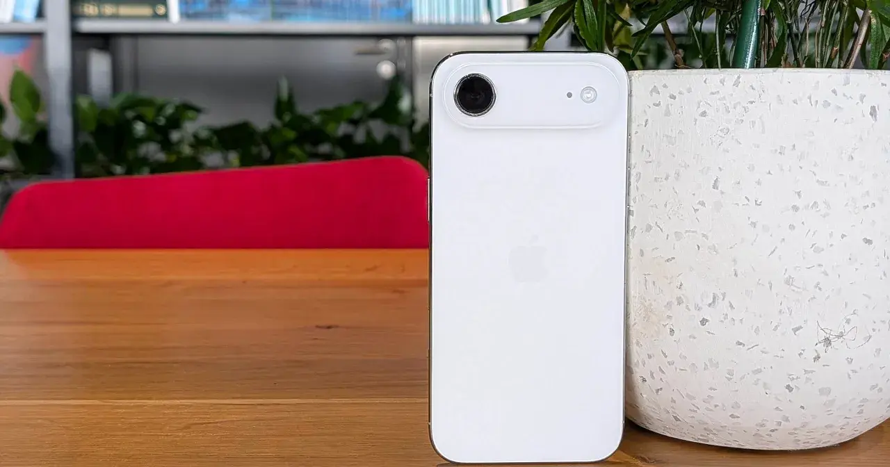 Biały iPhone 5c z charakterystycznym modułem aparatu, stojący na drewnianym blacie obok doniczki.