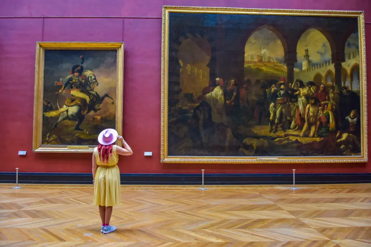 Kobieta w żółtej sukience i różowym kapeluszu podziwia obrazy w Luwrze. Co to za muzeum? To słynny paryski Luwr!