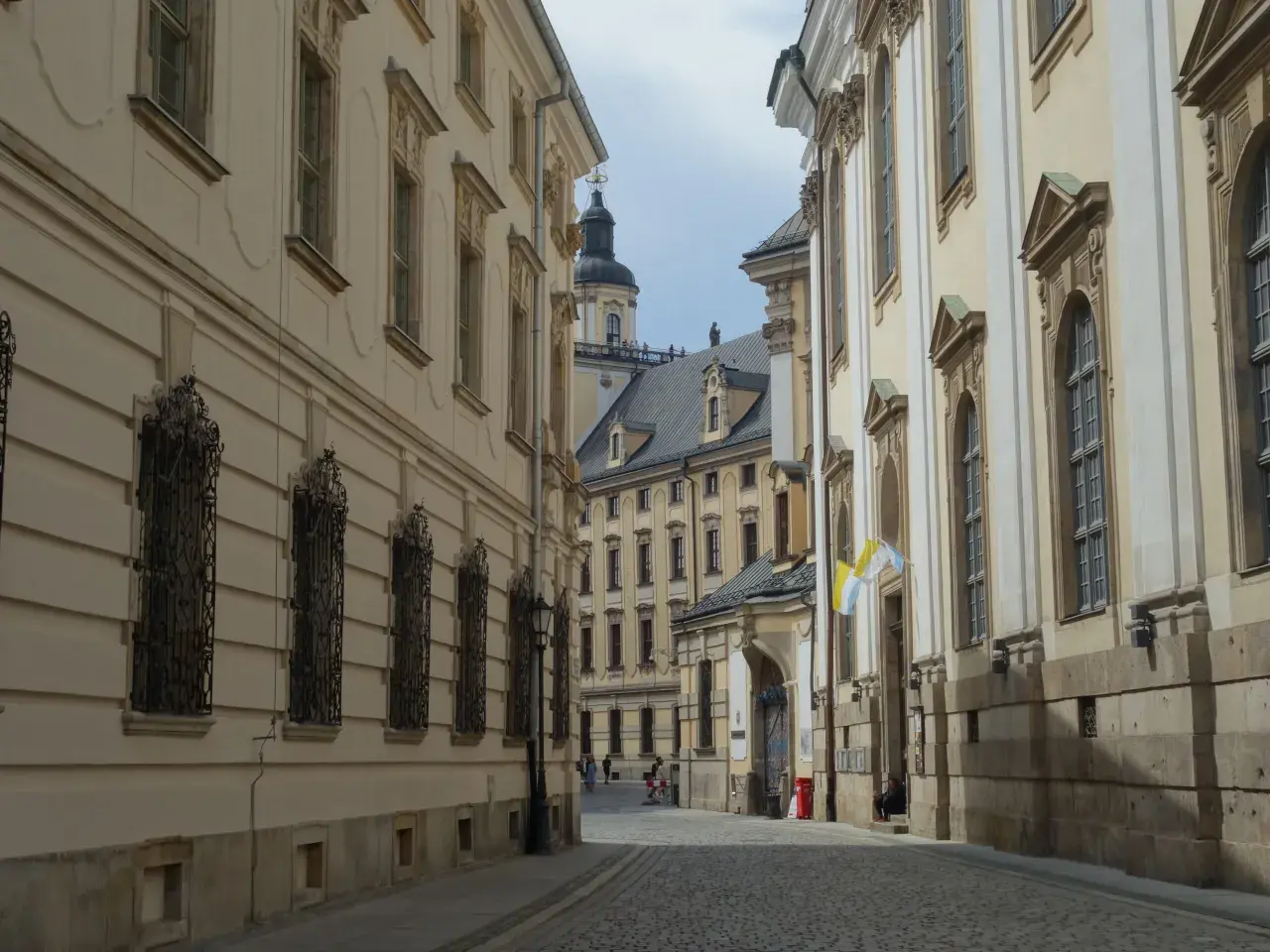 Wąska, brukowana uliczka prowadzi do serca starego rynku we Wrocławiu. Po bokach piętrzą się zabytkowe kamienice z ozdobnymi fasadami i kratami w oknach.