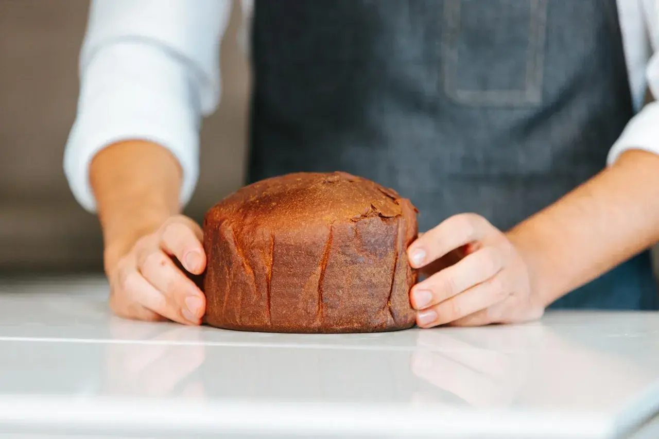Manos sostienen un pan brioche dorado y esponjoso sobre una superficie blanca.