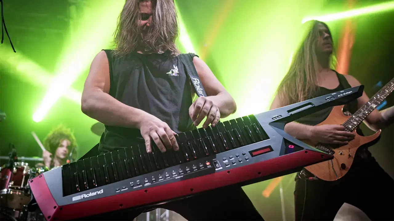 Muzyk gra na keyboardzie na pasku, podczas gdy gitarzysta i perkusista tworzą rockową atmosferę.