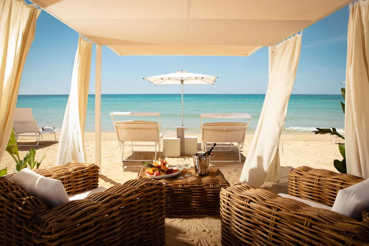 Lussuosa area lounge con divani in vimini, tavolino con frutta e champagne, lettini e ombrellone su una spiaggia di sabbia dorata. Un'oasi tra i migliori beach clubs Puglia.