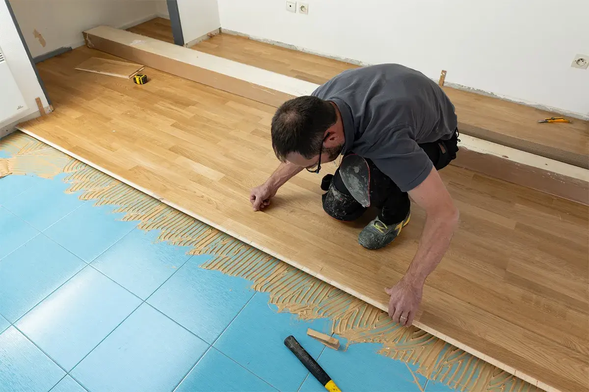 Pose d'un parquet stratifié sur un sol bleu carrelé, avec de la colle appliquée. L'homme ajuste une lame de bois.