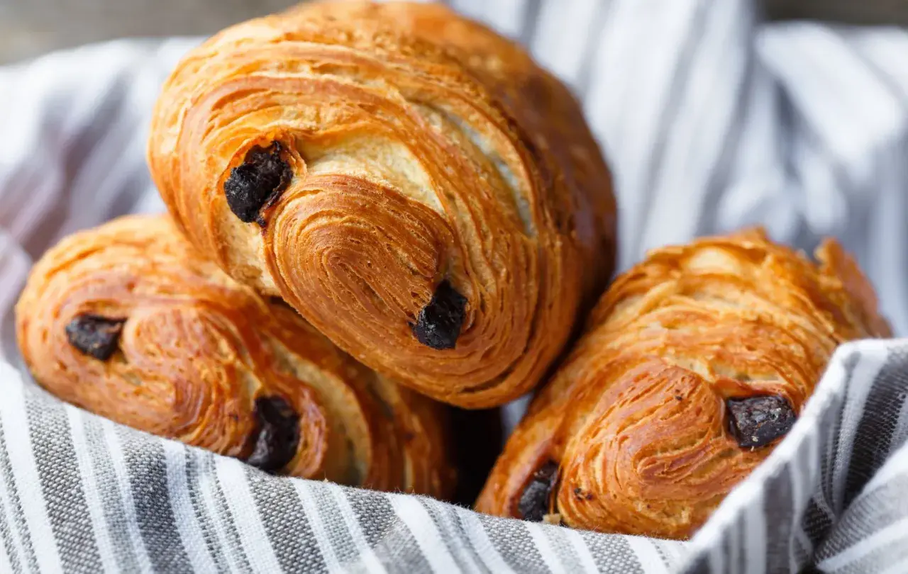 Tre deliziosi pain au chocolat dorati, con strati sfogliati e gocce di cioccolato fondente, adagiati su un canovaccio a righe.