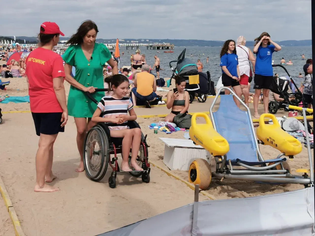 Na plaży dla niepełnosprawnych ratowniczka pomaga dziewczynce na wózku inwalidzkim. W tle widać innych plażowiczów i morze.