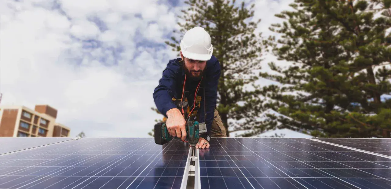 Técnico instala paneles solares, cumpliendo con el real decreto autoconsumo. Edificio y árboles al fondo.