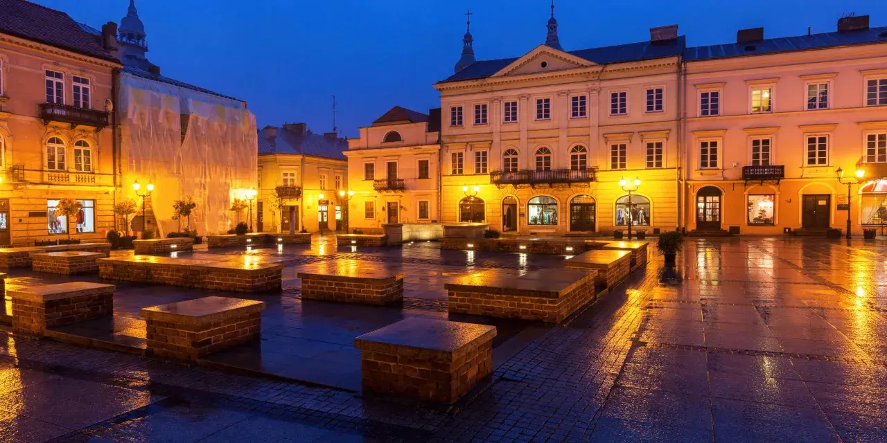 Piotrków Trybunalski atrakcje: Rynek nocą, mokry bruk odbija światła latarni i budynków. Widać kamienne ławki i fasady kamienic.