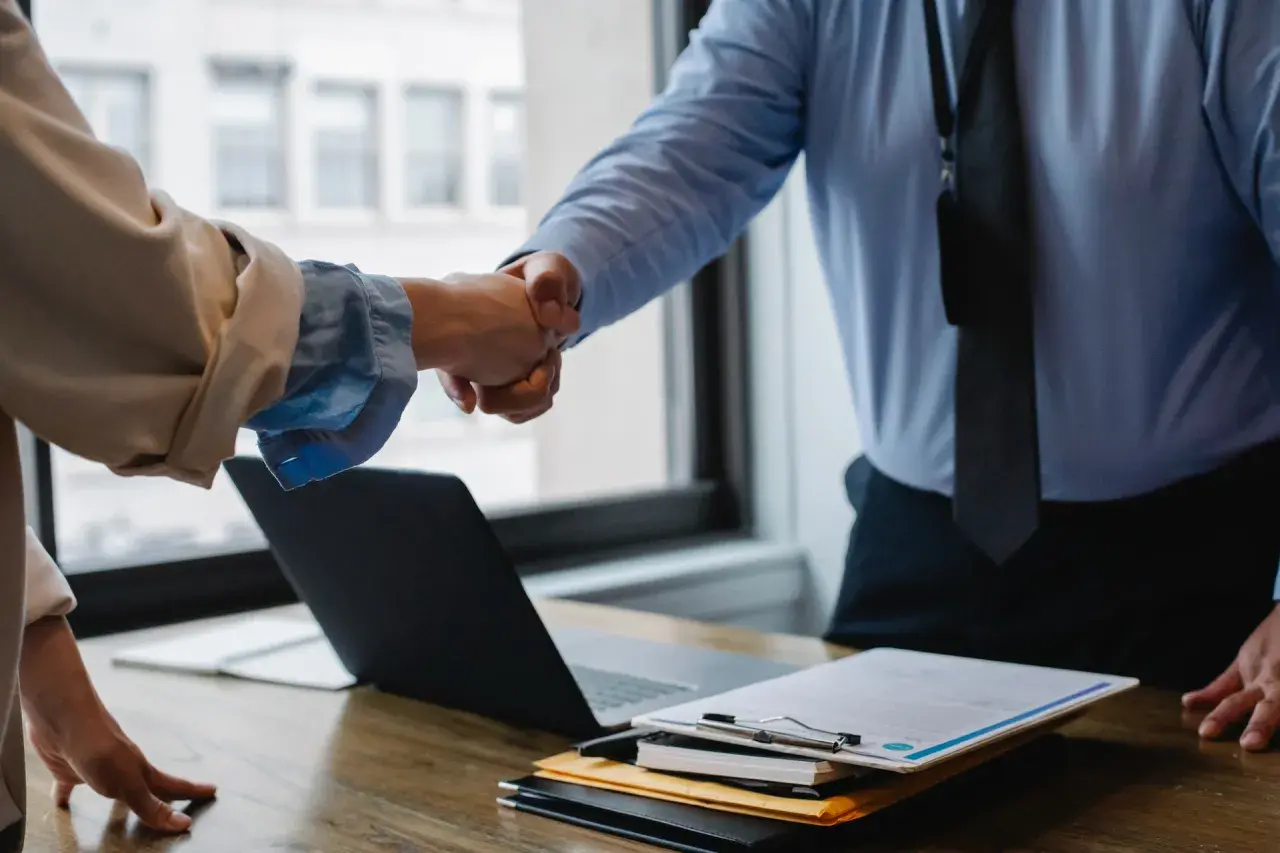 Dwie osoby ściskają sobie dłonie, symbolizując współpracę i inkluzywność w biznesie. Na stole leży laptop i dokumenty.