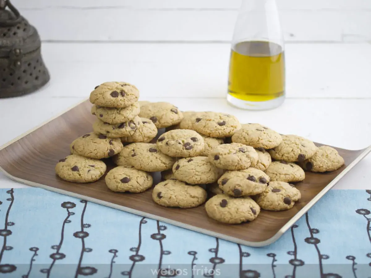 Montón de galletas caseras de pueblo con chispas de chocolate en una bandeja de madera, junto a una botella de aceite de oliva.