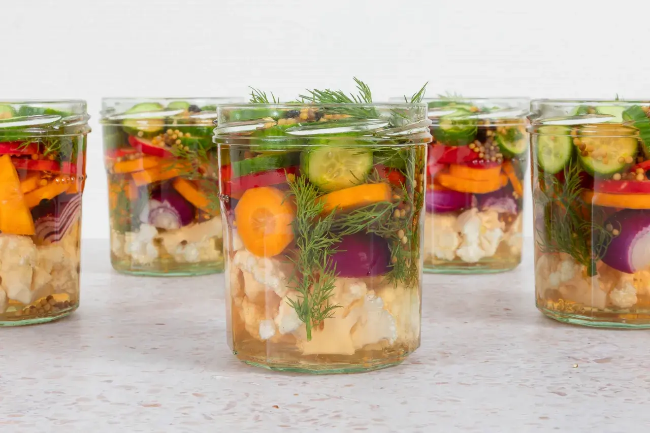 Sechs Gläser mit bunten Gemüsesorten wie Blumenkohl, Karotten, Gurken und Zwiebeln, bereit für mixed pickles selber machen.