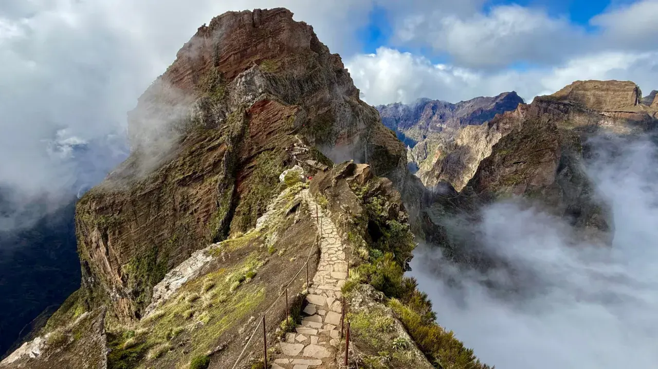 A stone path winds along a narrow mountain ridge, offering what is likely the best hiking in Portugal. Clouds drift through the dramatic, rugged peaks.