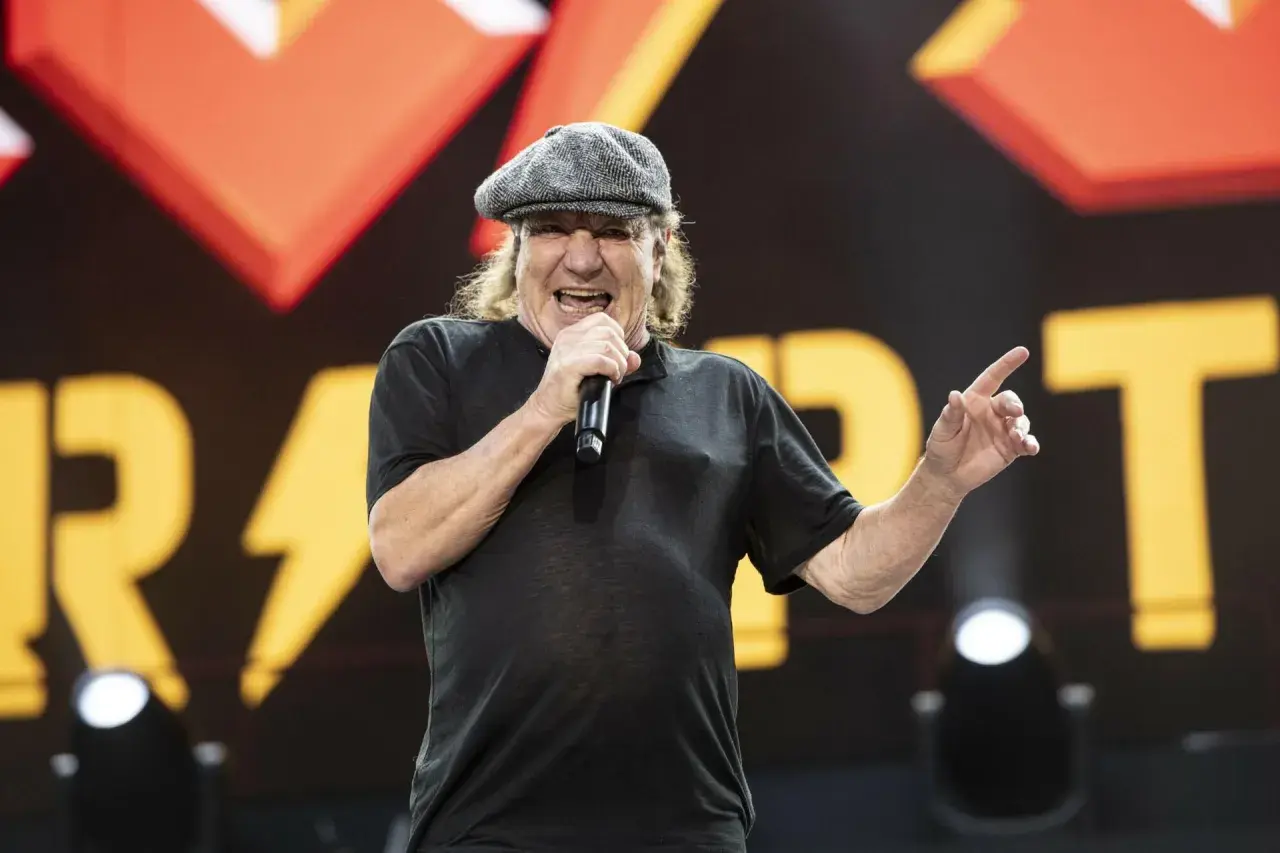 Brian Johnson young, w czapce i czarnej koszulce, śpiewa do mikrofonu na tle sceny z logo zespołu.