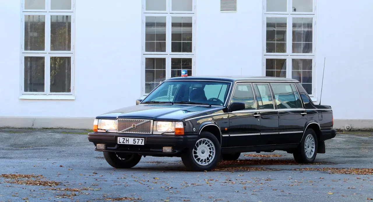 Svart, förlängd 760 Volvo med blåljus på taket, parkerad framför en vit byggnad med stora fönster.