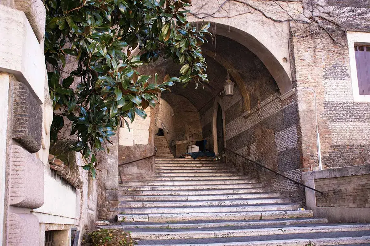 Scalinata romana con alberi di magnolia e un arco antico. Un'idea per cosa vedere a Roma domani, un angolo suggestivo e storico.