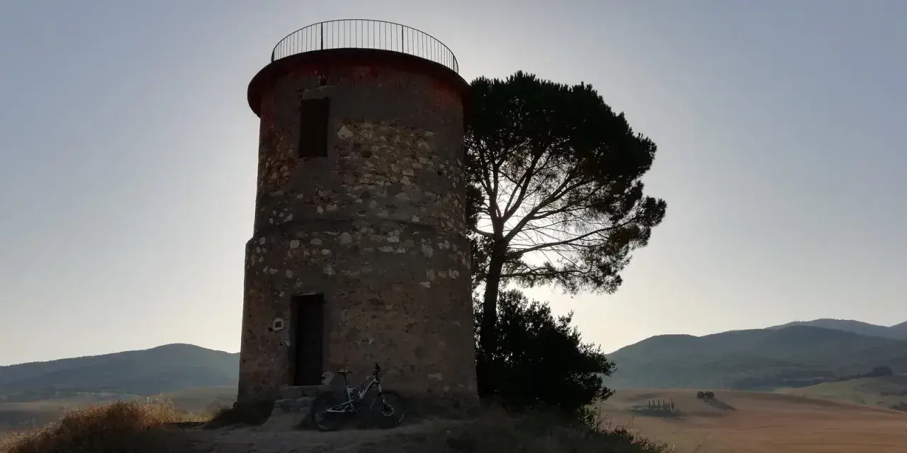 Torre circolare in pietra con una bicicletta appoggiata, vicina a un pino solitario. Paesaggio collinare sullo sfondo, forse la macchia della Magona.