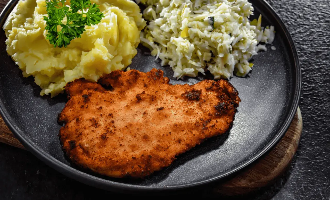 Kotlet schabowy z ziemniakami i surówką na ciemnym talerzu.