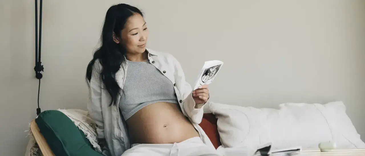 Gravid kvinna tittar på ultraljudsbild, i trimester 2. Hon sitter bekvämt i soffan med sin mage synlig.