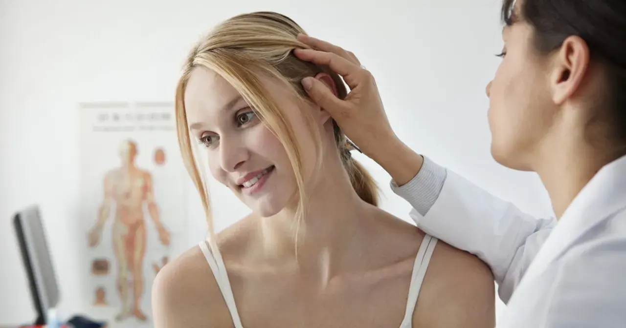Dermatolog bada skórę głowy młodej kobiety. W tle wisi plakat anatomiczny.