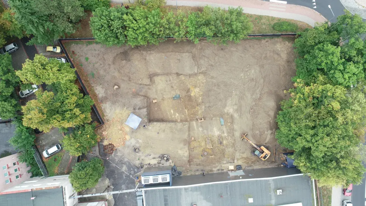 Wykop archeologiczny na placu budowy, gdzie prowadzone są badania przed pozwoleniem na budowę. Widoczny koparka i ślady dawnej zabudowy.