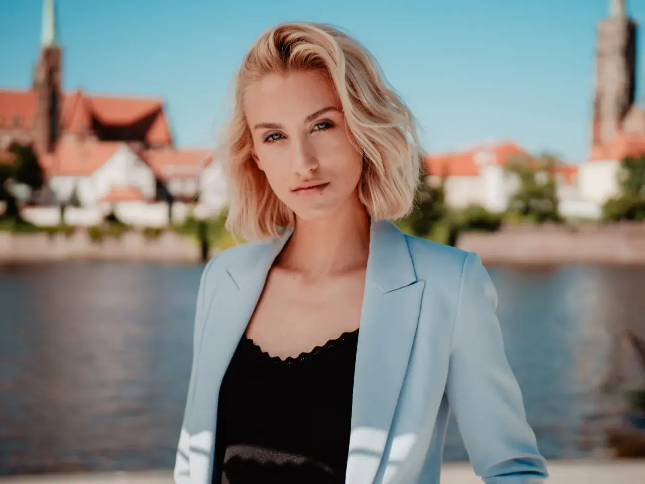 Portret fotograficzny młodej kobiety z blond włosami, w jasnoniebieskiej marynarce, na tle miasta i rzeki.