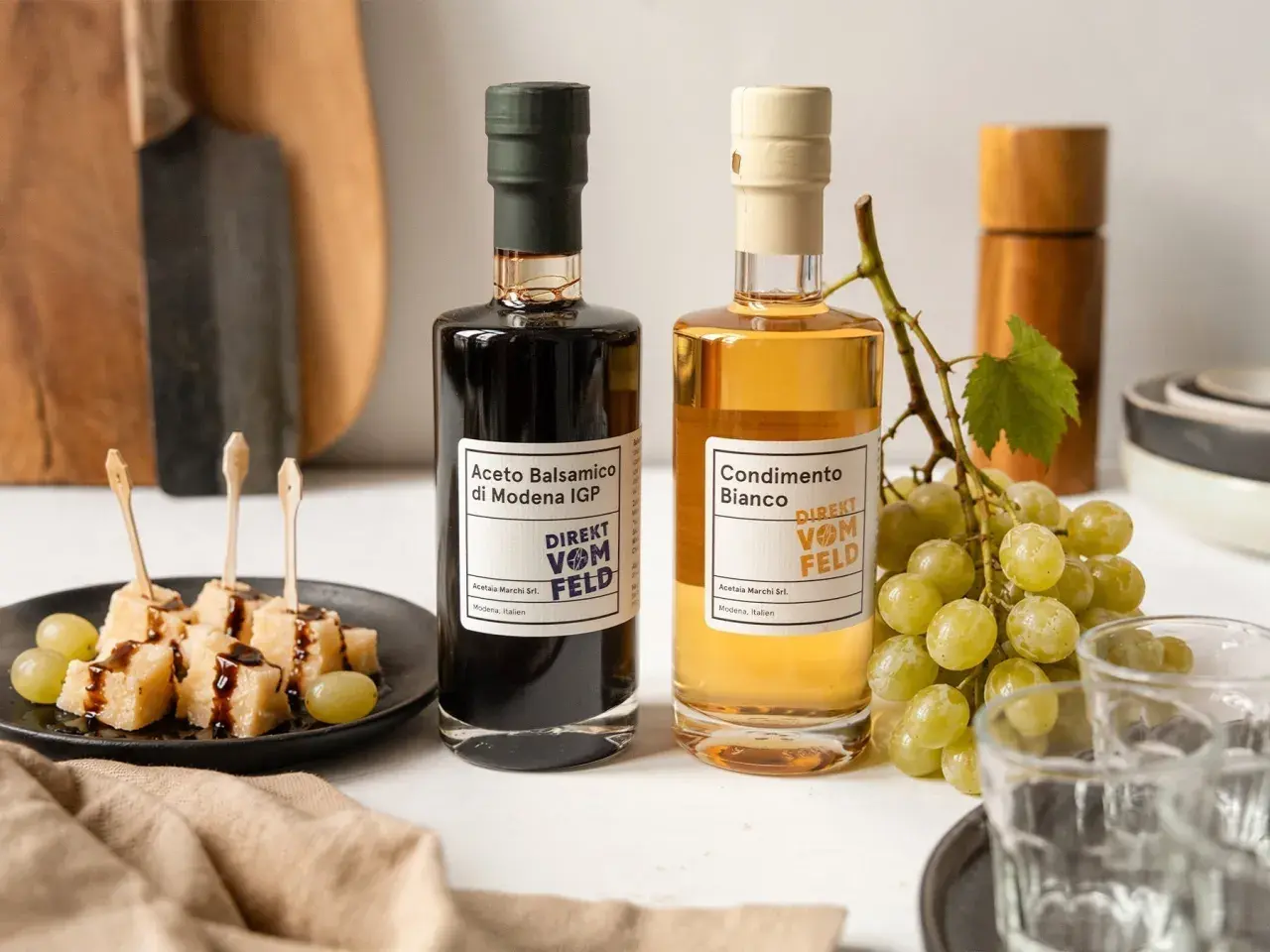 Zwei Flaschen Balsamico und Condimento Bianco, daneben Käsewürfel mit Balsamico und Trauben.
