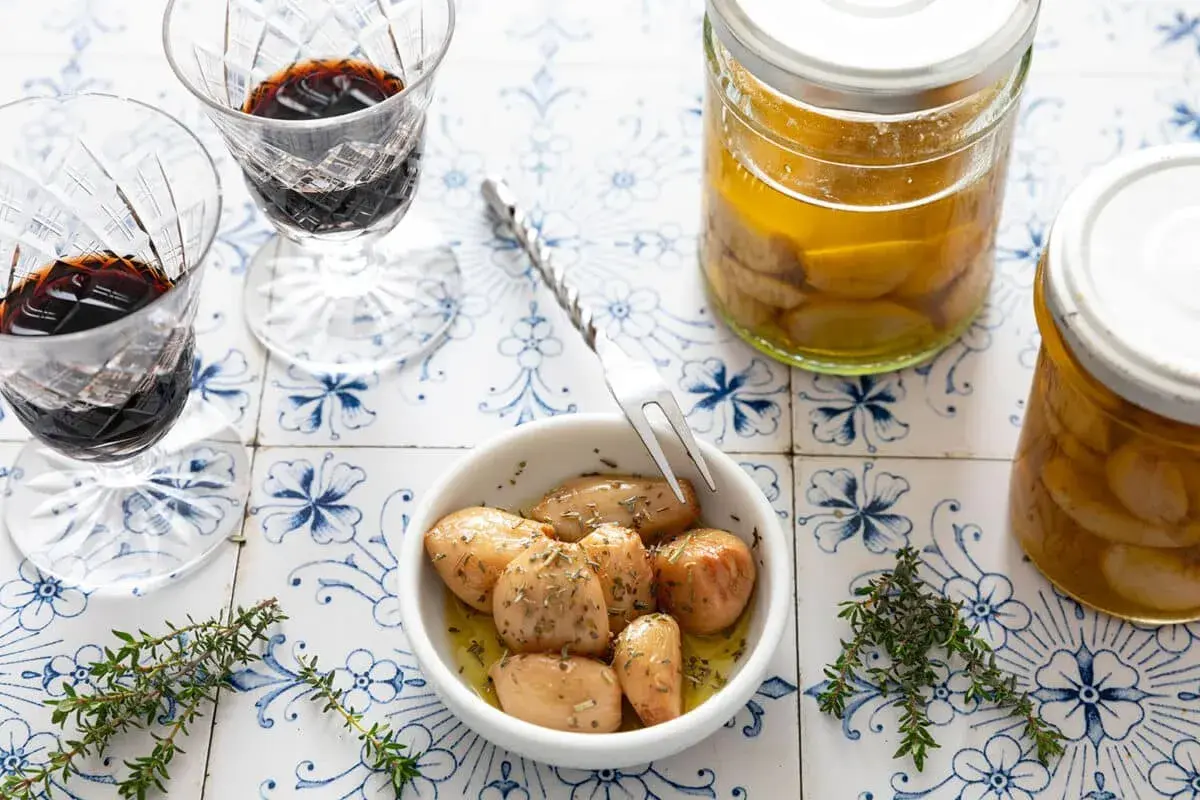 Ail confit au four, tendre et parfumé, servi dans un bol avec des herbes. Deux verres de vin rouge et des bocaux d'ail confit complètent la scène.
