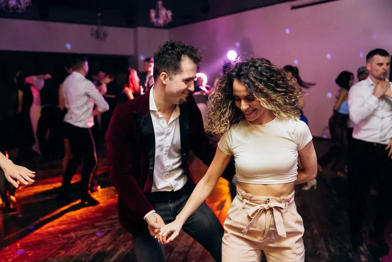 Para tańczy w rytm muzyki, ciesząc się wspólnym tańcem użytkowym. W tle widać innych bawiących się ludzi.
