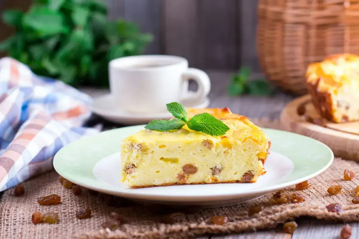 Una fetta di torta putana, soffice e dorata, con uvetta e una fogliolina di menta, accanto a una tazza di caffè.