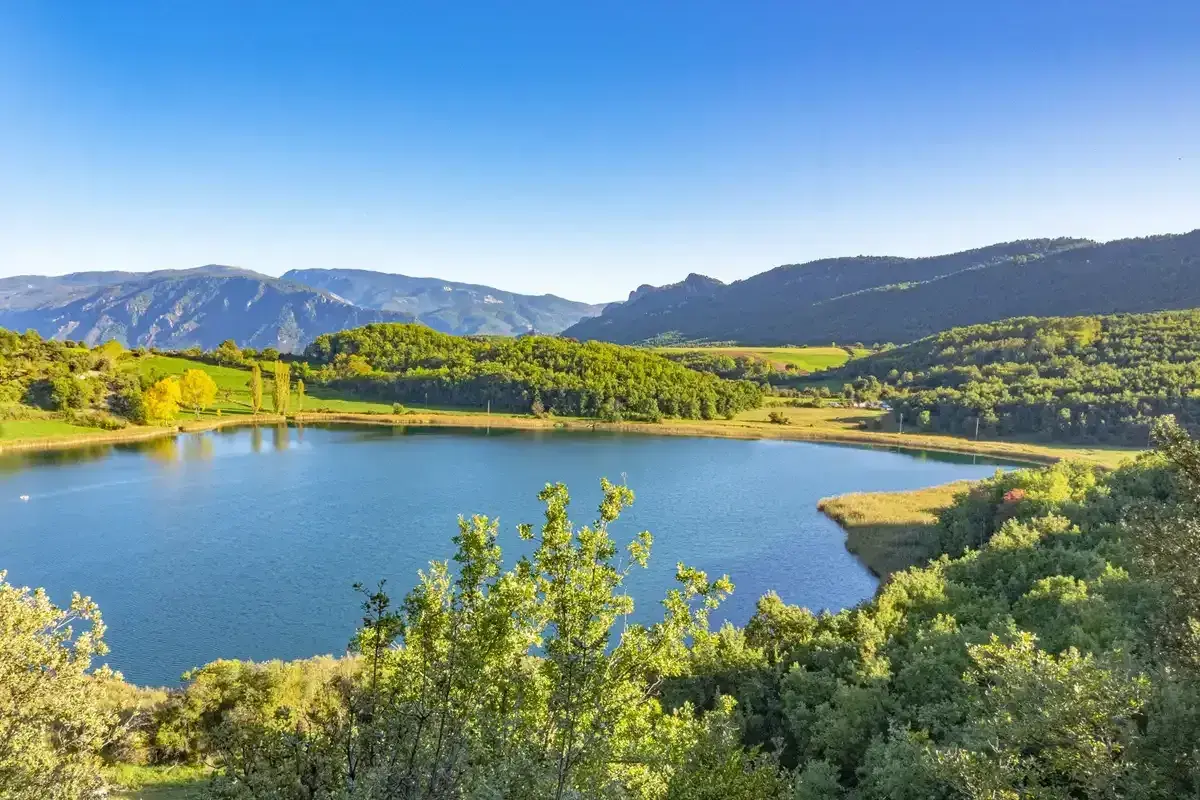 Paisaje sereno en la conca de dalt: un lago azul rodeado de colinas verdes y montañas bajo un cielo despejado.