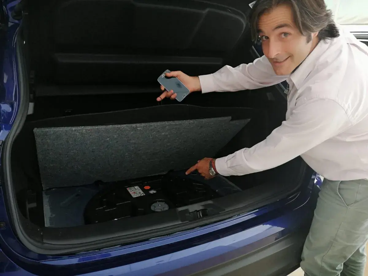 Hombre señala el espacio vacío en el maletero de un coche azul, donde debería estar la rueda de repuesto. "He pinchado y no tengo rueda de repuesto", parece decir.