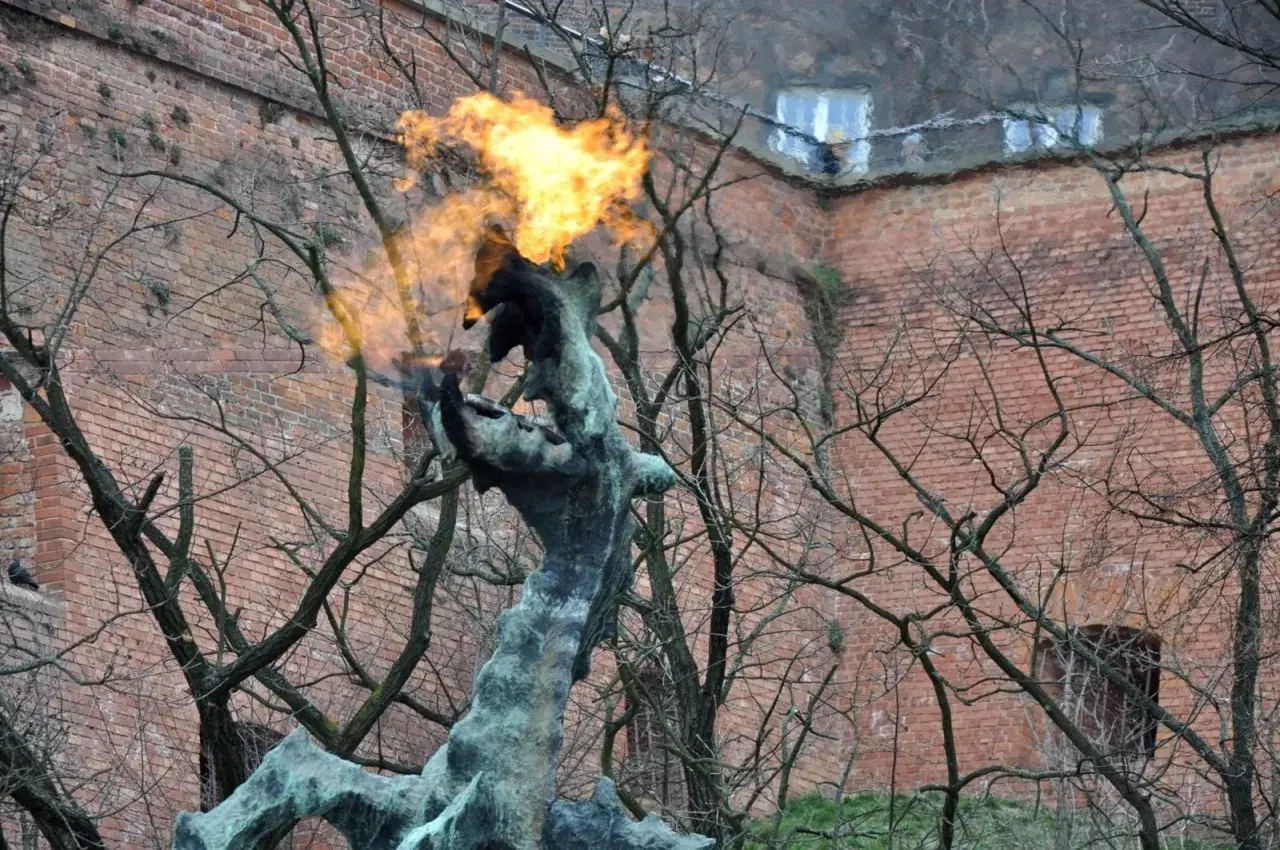 Pomnik smoka wawelskiego zieje ogniem na tle ceglanej ściany.