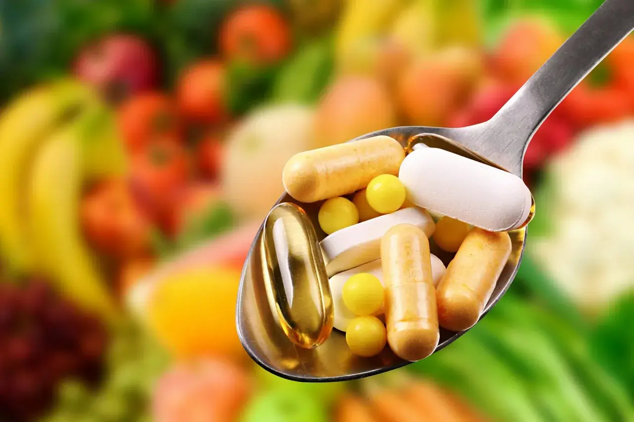 Löffel voller Pillen und Kapseln, die bei Krankheit helfen. Welche Vitamine wenn man krank ist? Bunte Früchte im Hintergrund.