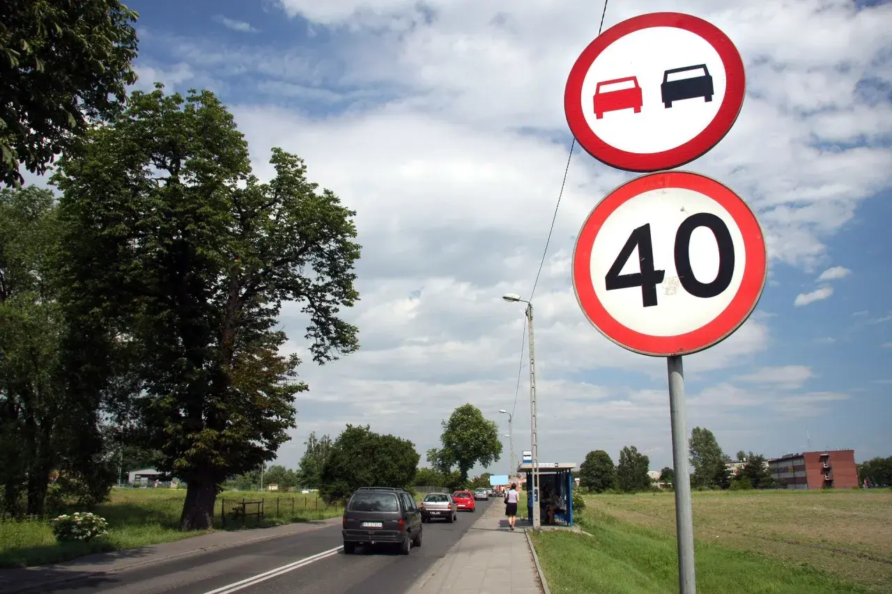 Znaki drogowe: zakaz wyprzedzania i ograniczenie prędkości do 40 km/h. Samochody jadą drogą, po prawej stronie przystanek autobusowy.