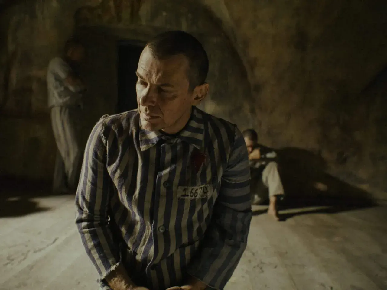 Film o Maksymilianie Kolbe: więźniowie w pasiakach, jeden z nich z numerem 16670, w ponurym wnętrzu.