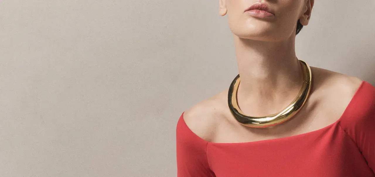 A woman wears a bold gold necklace with a curved neckline, complementing her red off-the-shoulder top.