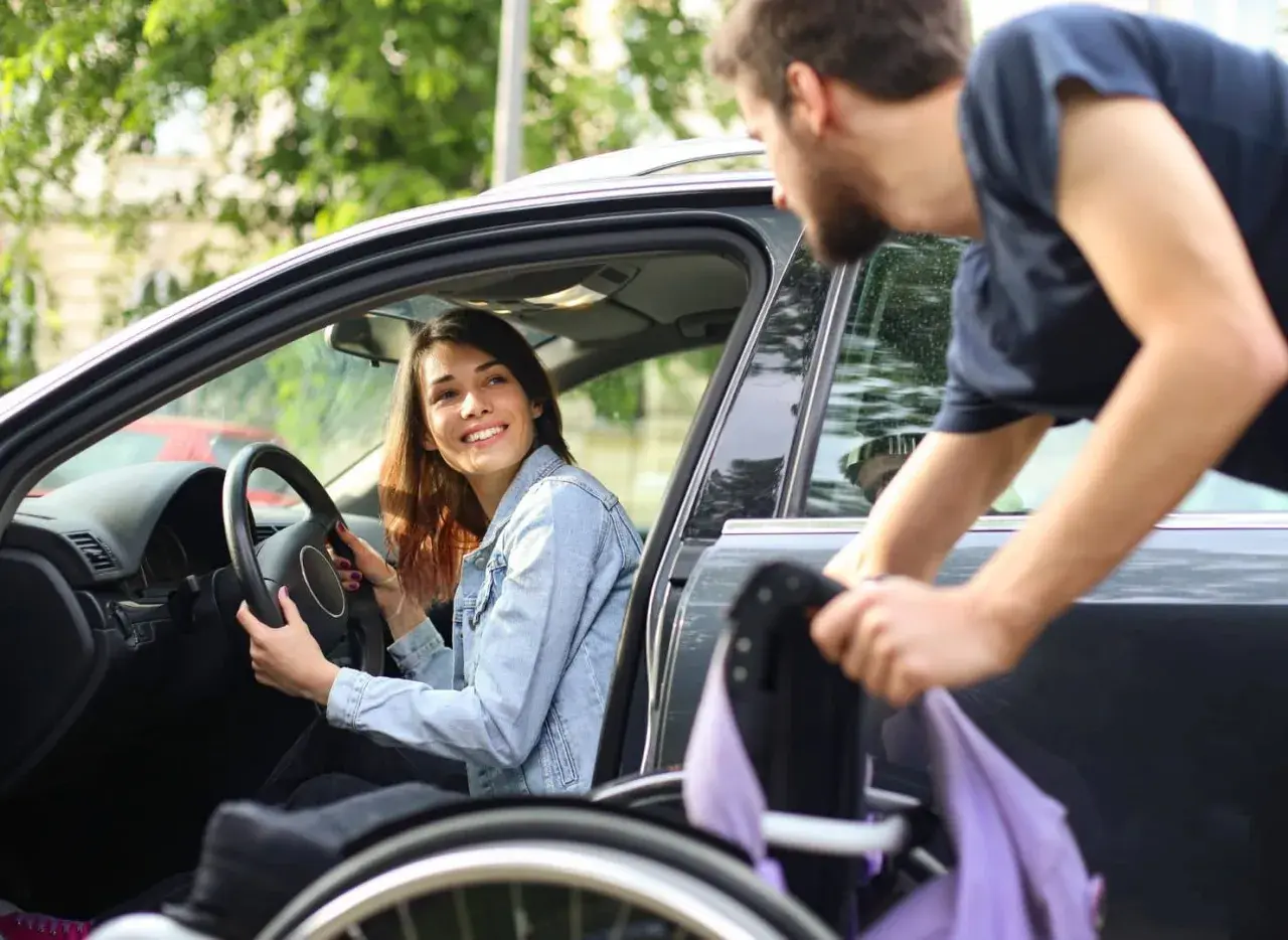 Una donna disabile sorride al volante mentre un uomo aiuta a caricare la sua sedia a rotelle in auto.