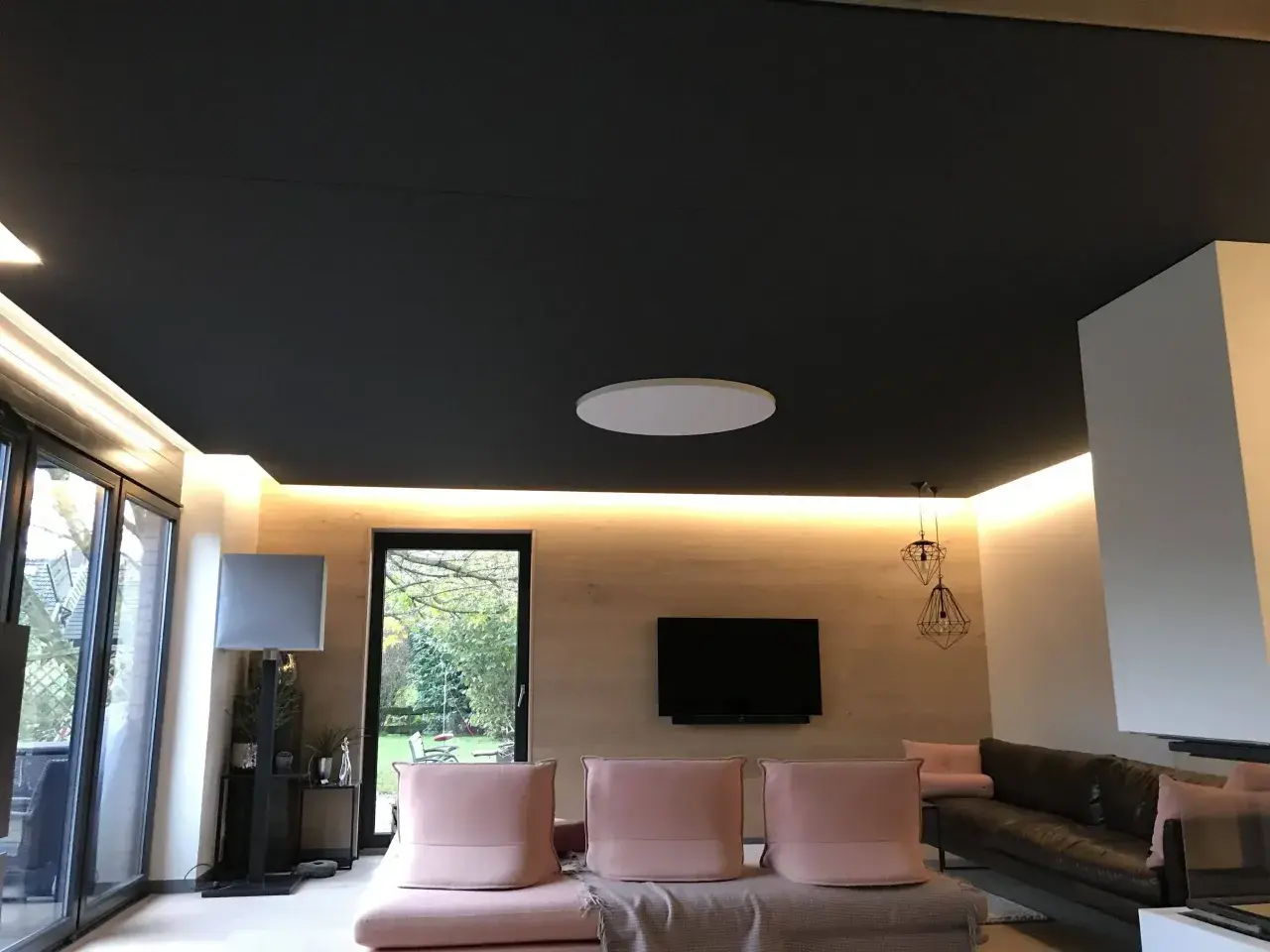 Ciemny sufit napinany z subtelnym oświetleniem LED. W salonie różowe kanapy, czarna skórzana sofa i telewizor.