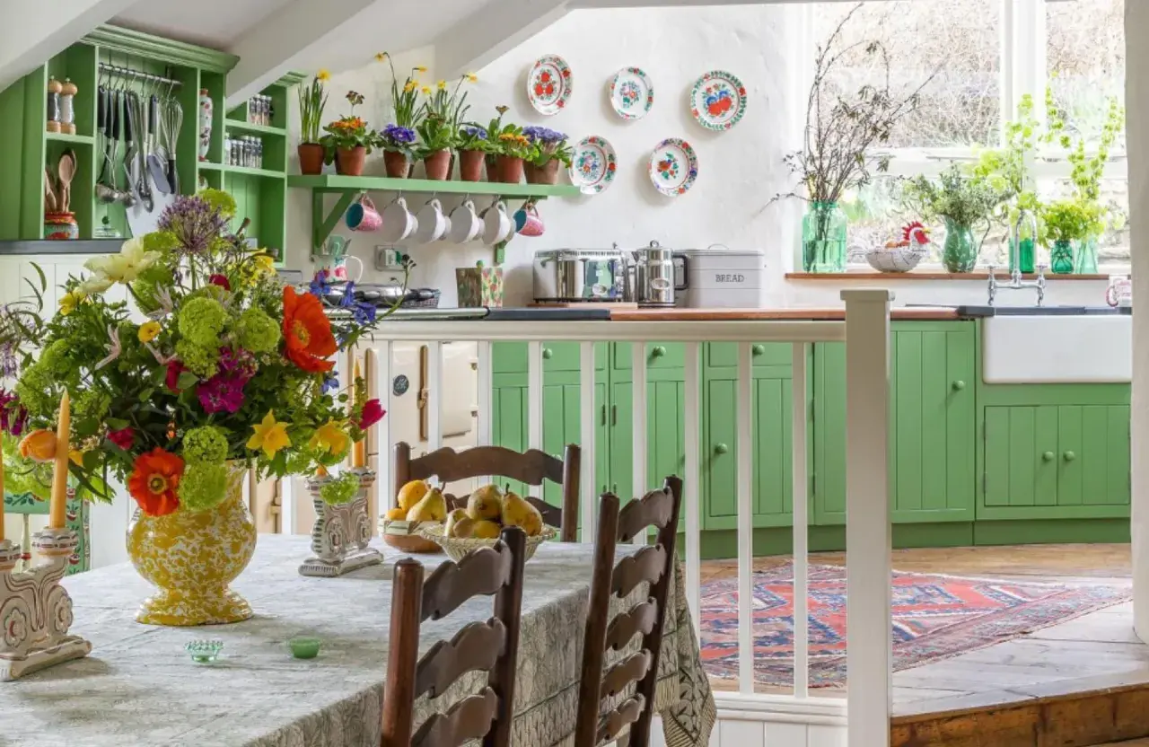 Une cuisine lumineuse avec des armoires vertes vives, un grand bouquet de fleurs colorées sur la table, et une décoration style cottage ou campagne chic.